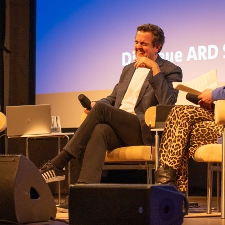 Bastian Pastewka sitzt mit 2 Moderatorinnen lachend auf der Bühne